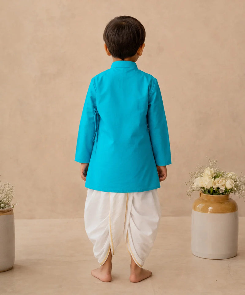 A child viewed from the back wearing a bright turquoise-blue kurta and white dhoti pants, standing barefoot against a warm beige textured wall with decorative ceramic vases on either side.
