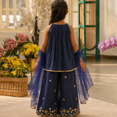 Woman in a navy blue traditional outfit with floral embroidery standing in a decorated room with flowers.