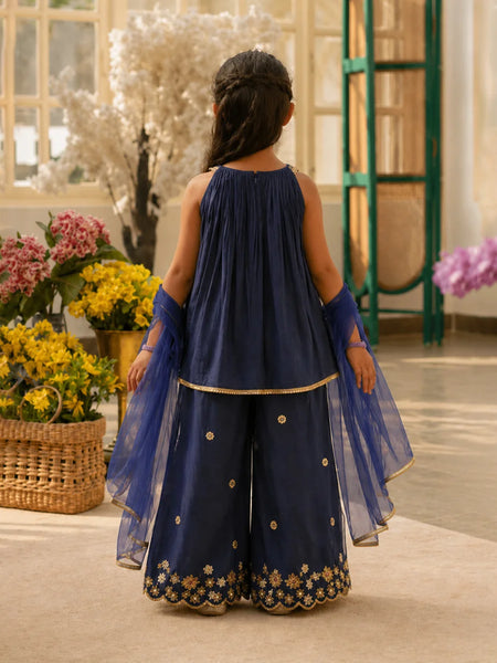 Woman in a navy blue traditional outfit with floral embroidery standing in a decorated room with flowers.