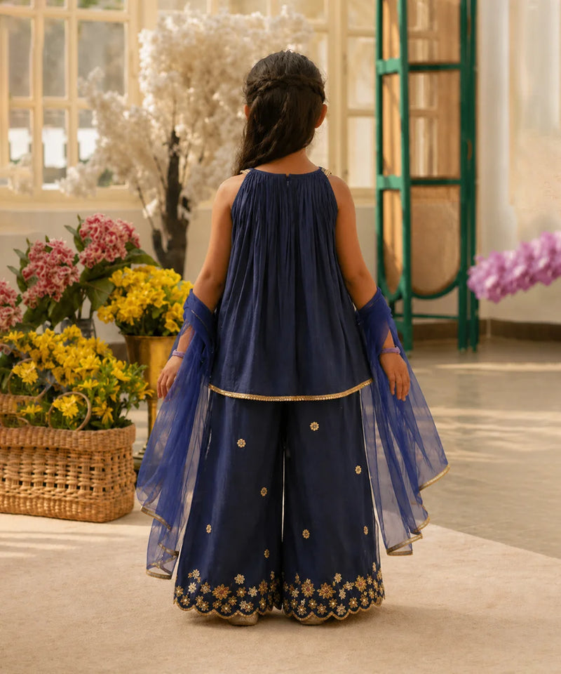 Woman in a navy blue traditional outfit with floral embroidery standing in a decorated room with flowers.