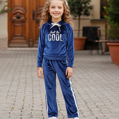 A young girl with curly hair stands on a paved outdoor walkway wearing a royal blue velour tracksuit with white side stripes and a white drawstring. The hoodie features the text 'Just Be COOL' in white and silver.