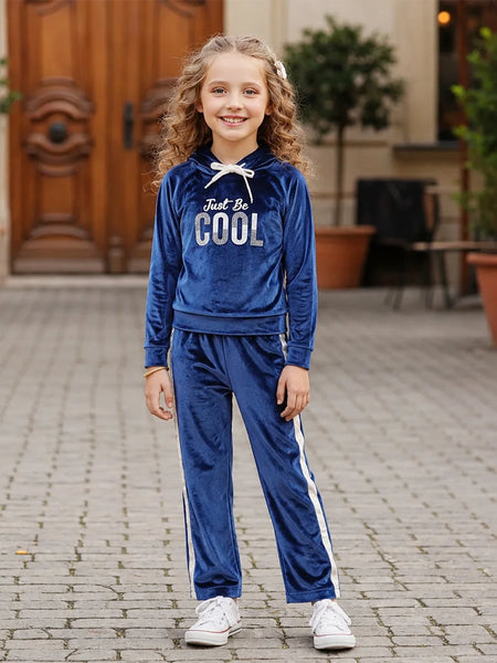 A young girl with curly hair stands on a paved outdoor walkway wearing a royal blue velour tracksuit with white side stripes and a white drawstring. The hoodie features the text 'Just Be COOL' in white and silver.