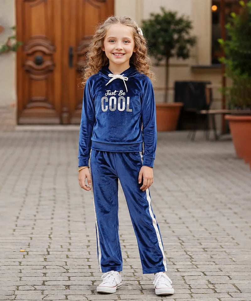 A young girl with curly hair stands on a paved outdoor walkway wearing a royal blue velour tracksuit with white side stripes and a white drawstring. The hoodie features the text 'Just Be COOL' in white and silver.