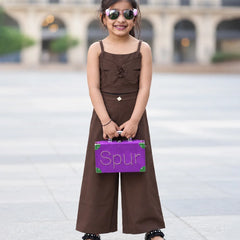 A young girl stands outdoors in a plaza wearing a brown sleeveless wide-leg jumpsuit, holding a purple suitcase with the word 'Spur' on the front, and wearing sunglasses.