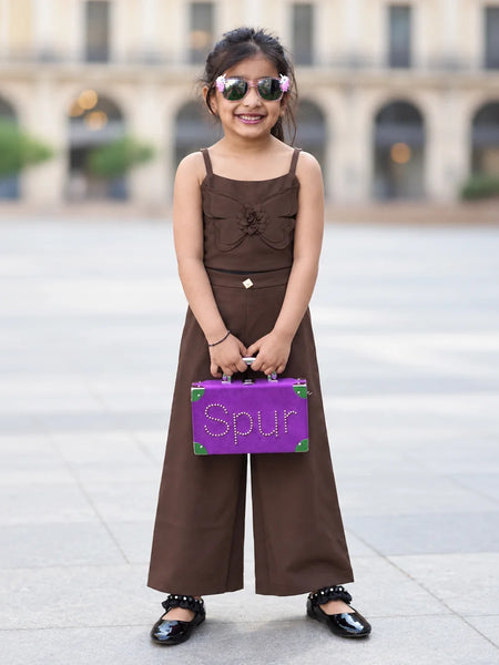 A young girl stands outdoors in a plaza wearing a brown sleeveless wide-leg jumpsuit, holding a purple suitcase with the word 'Spur' on the front, and wearing sunglasses.