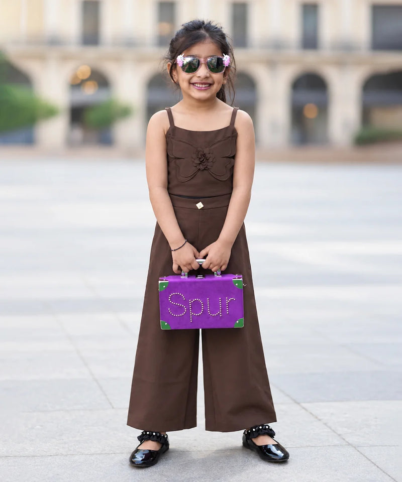 A young girl stands outdoors in a plaza wearing a brown sleeveless wide-leg jumpsuit, holding a purple suitcase with the word 'Spur' on the front, and wearing sunglasses.