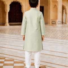 A young boy viewed from behind, wearing a light sage green kurta and white trousers, standing in a grand, historic courtyard with beige stone arches and tiled floors.
