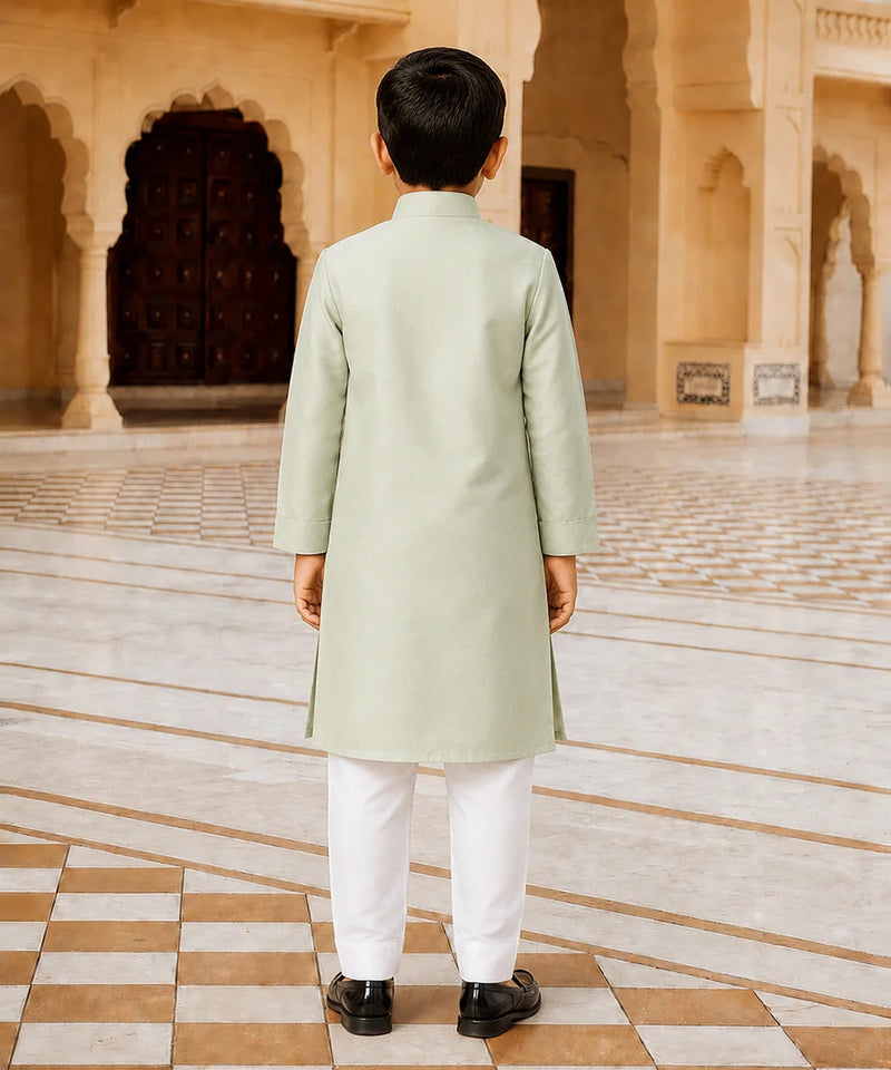 A young boy viewed from behind, wearing a light sage green kurta and white trousers, standing in a grand, historic courtyard with beige stone arches and tiled floors.