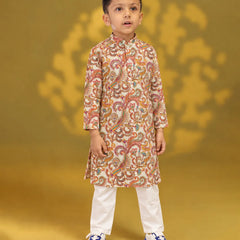 A young boy modeling a traditional Indian outfit consisting of a beige long-sleeve kurta with a colorful paisley pattern, paired with white trousers and blue sneakers, set against a golden-yellow background.