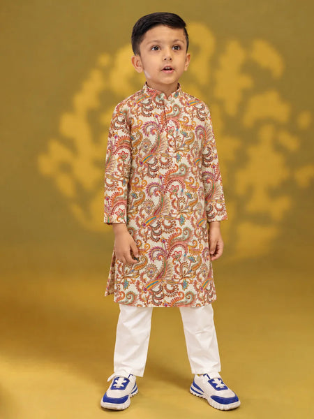 A young boy modeling a traditional Indian outfit consisting of a beige long-sleeve kurta with a colorful paisley pattern, paired with white trousers and blue sneakers, set against a golden-yellow background.