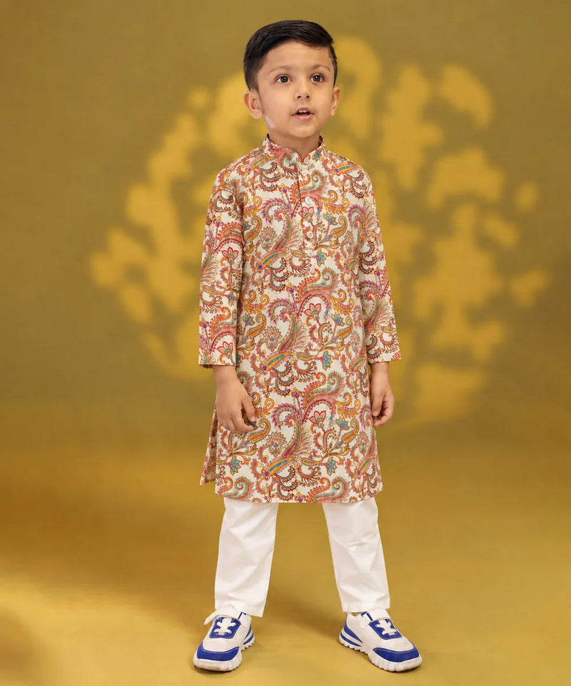 A young boy modeling a traditional Indian outfit consisting of a beige long-sleeve kurta with a colorful paisley pattern, paired with white trousers and blue sneakers, set against a golden-yellow background.