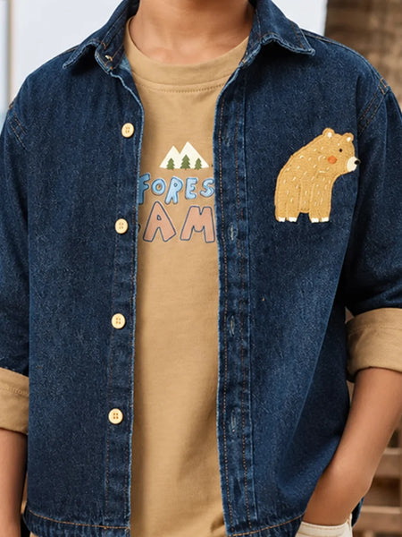 A child wearing a dark blue denim button-up shirt over a tan graphic t-shirt. The denim shirt features rolled cuffs, gold-tone buttons, and a brown bear patch on the chest.