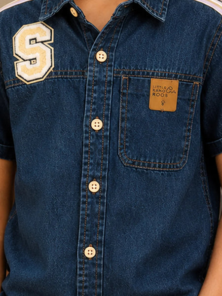 Close-up of a blue denim short-sleeve button-up shirt with a large cream letter 'S' patch on the left chest, a brown leather patch on the right chest pocket, and cream buttons.