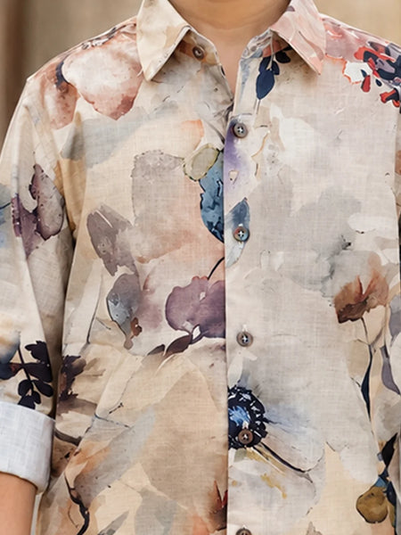 A close-up of a men's long-sleeve button-up shirt featuring a watercolor floral print on a beige linen-like base. The print includes soft shades of brown, purple, blue, and red, with the sleeves rolled up to show a light gray undershirt.