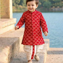 Child wearing a red traditional outfit with white patterns, standing by a body of water.