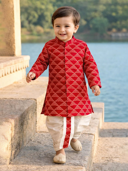 Child wearing a red traditional outfit with white patterns, standing by a body of water.