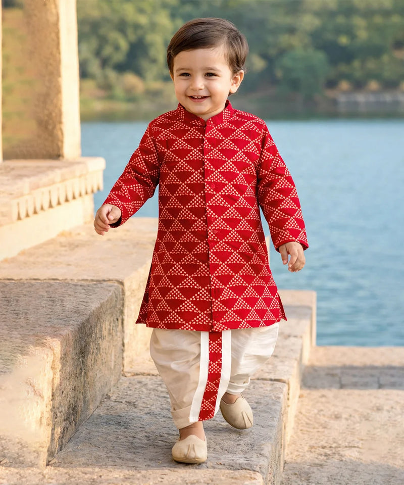 Child wearing a red traditional outfit with white patterns, standing by a body of water.