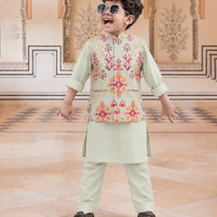 A young boy wearing a light sage green traditional Indian outfit with a colorful embroidered vest, matching trousers, and sunglasses, posing in an ornate indoor setting.