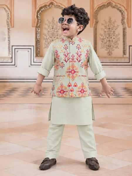 A young boy wearing a light sage green traditional Indian outfit with a colorful embroidered vest, matching trousers, and sunglasses, posing in an ornate indoor setting.