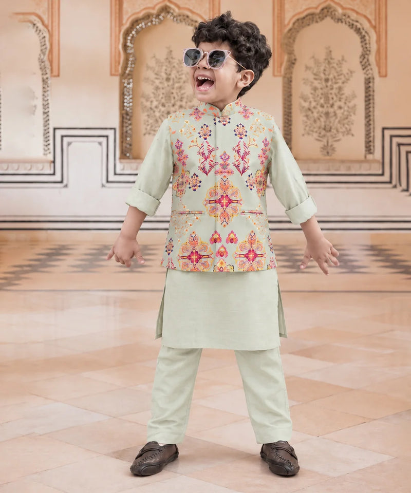 A young boy wearing a light sage green traditional Indian outfit with a colorful embroidered vest, matching trousers, and sunglasses, posing in an ornate indoor setting.