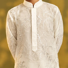 A close-up of a child wearing a cream-colored embroidered kurta with silver sequin detailing, set against a soft, blurred golden-yellow background.