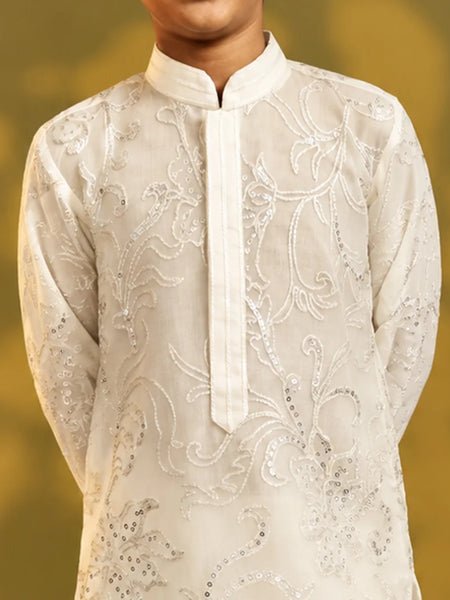A close-up of a child wearing a cream-colored embroidered kurta with silver sequin detailing, set against a soft, blurred golden-yellow background.