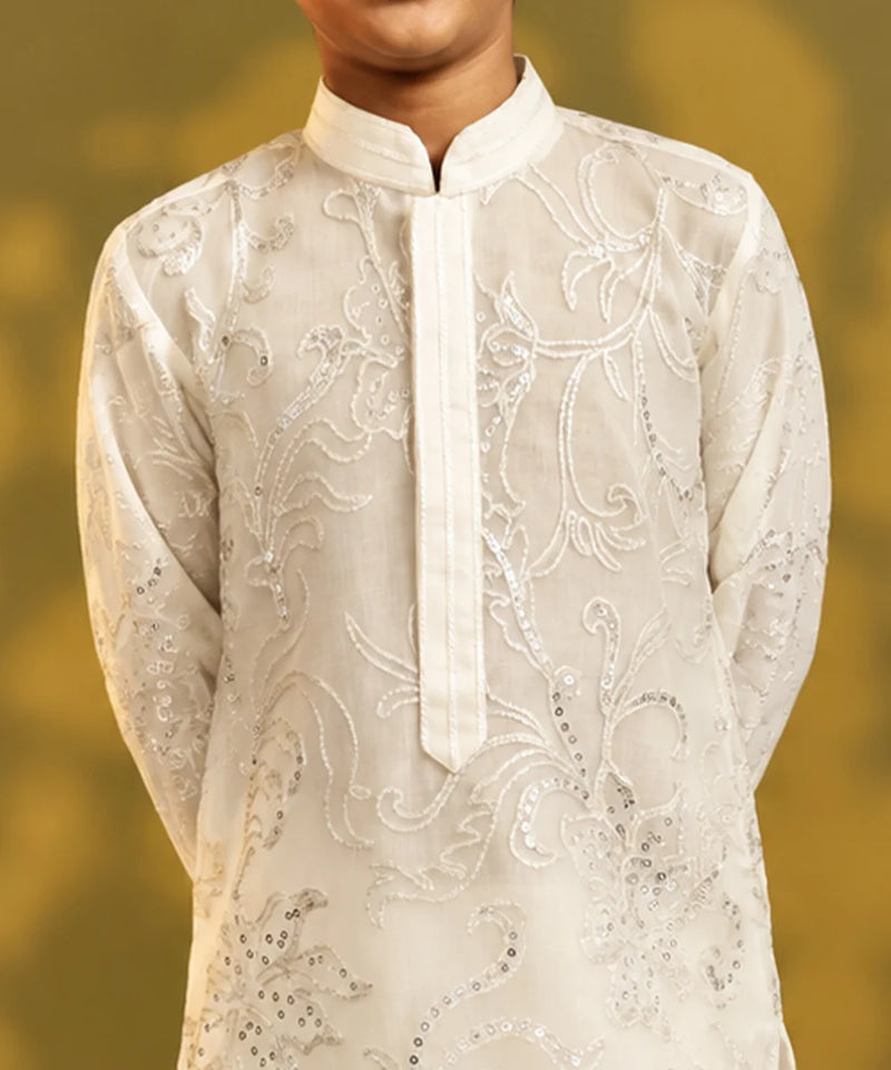 A close-up of a child wearing a cream-colored embroidered kurta with silver sequin detailing, set against a soft, blurred golden-yellow background.