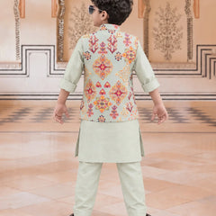A child viewed from behind wearing a light sage green traditional outfit with a sleeveless vest featuring a colorful geometric and floral pattern in orange, red, and yellow. The child is standing in a grand hall with beige walls and patterned tile floors.