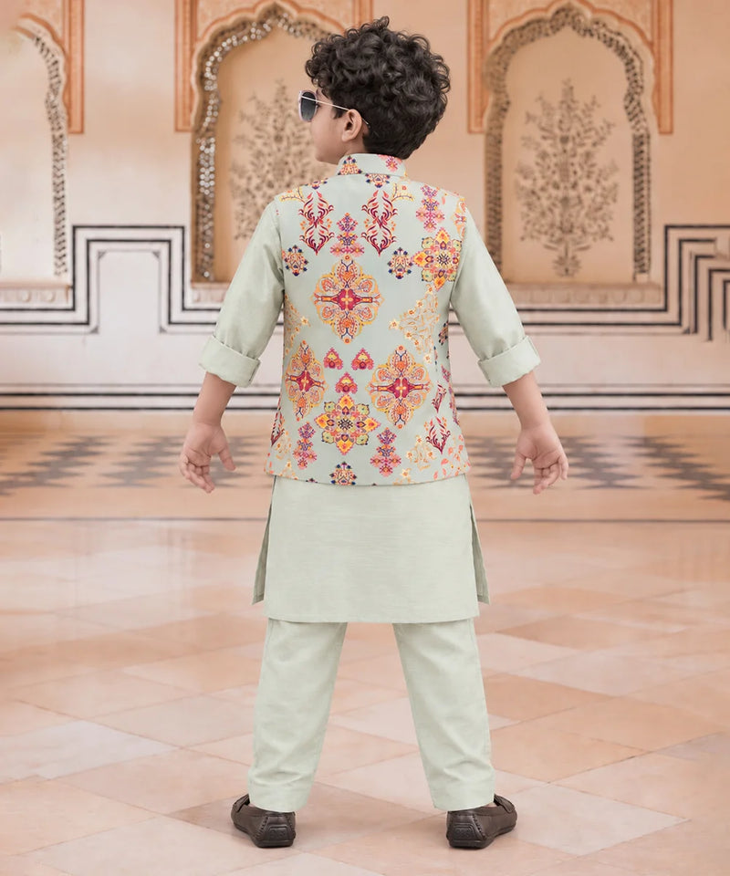 A child viewed from behind wearing a light sage green traditional outfit with a sleeveless vest featuring a colorful geometric and floral pattern in orange, red, and yellow. The child is standing in a grand hall with beige walls and patterned tile floors.