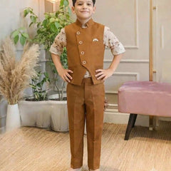 A child viewed from behind wearing a brown vest and matching trousers over a white patterned short-sleeve shirt, standing in an indoor room with plants, a lamp, and a pink bench.