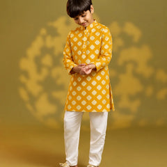 A child viewed from behind wearing a mustard yellow kurta with a white geometric diamond pattern, paired with white trousers and sandals, standing against a warm yellow background with soft shadow details.