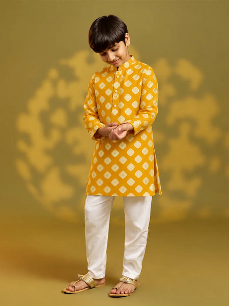 A child viewed from behind wearing a mustard yellow kurta with a white geometric diamond pattern, paired with white trousers and sandals, standing against a warm yellow background with soft shadow details.