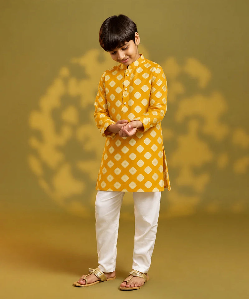 A child viewed from behind wearing a mustard yellow kurta with a white geometric diamond pattern, paired with white trousers and sandals, standing against a warm yellow background with soft shadow details.