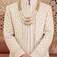 A close-up of a person wearing a cream-colored embroidered sherwani with a mandarin collar and layered gold and pearl necklaces, standing in front of a patterned wooden screen.
