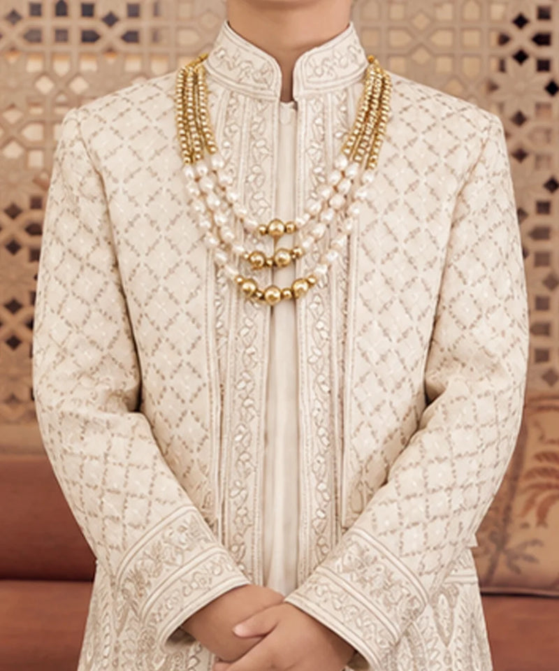 A close-up of a person wearing a cream-colored embroidered sherwani with a mandarin collar and layered gold and pearl necklaces, standing in front of a patterned wooden screen.