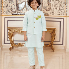 A young boy wearing a light mint green traditional Indian outfit with a small embroidered motif on the chest, standing in front of an ornate gold console table and a decorative wall.
