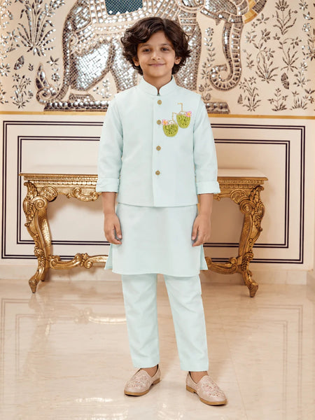 A young boy wearing a light mint green traditional Indian outfit with a small embroidered motif on the chest, standing in front of an ornate gold console table and a decorative wall.