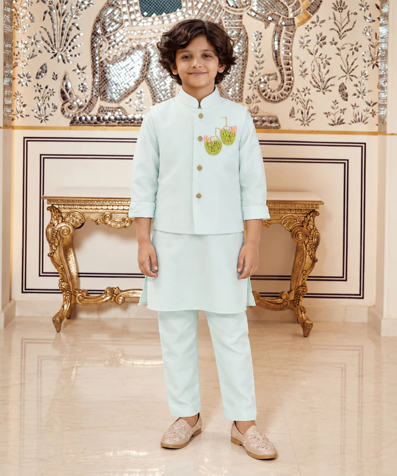 A young boy wearing a light mint green traditional Indian outfit with a small embroidered motif on the chest, standing in front of an ornate gold console table and a decorative wall.