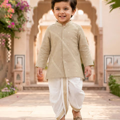 A young boy smiling and walking outdoors in a beige embroidered kurta with white dhoti pants and metallic shoes, set against a warm architectural background with arches and flowers.