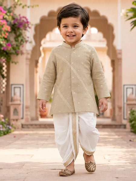 A young boy smiling and walking outdoors in a beige embroidered kurta with white dhoti pants and metallic shoes, set against a warm architectural background with arches and flowers.