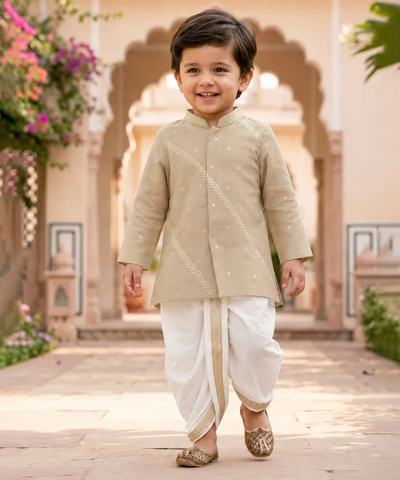 A young boy smiling and walking outdoors in a beige embroidered kurta with white dhoti pants and metallic shoes, set against a warm architectural background with arches and flowers.
