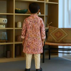 Child wearing a pink floral kurta standing in a room with a bookshelf and chair.