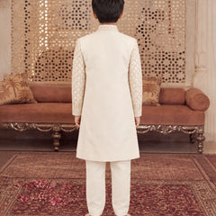A child viewed from behind wearing a cream-colored traditional outfit with patterned sleeves, standing on a patterned rug in front of an ornate brown sofa and decorative wall panel.