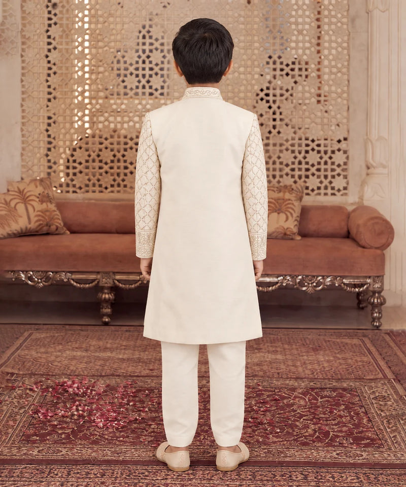 A child viewed from behind wearing a cream-colored traditional outfit with patterned sleeves, standing on a patterned rug in front of an ornate brown sofa and decorative wall panel.