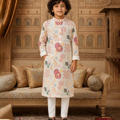 A young boy stands in a decorated traditional room wearing a cream-colored kurta with colorful geometric and floral patterns, paired with white trousers and black loafers.