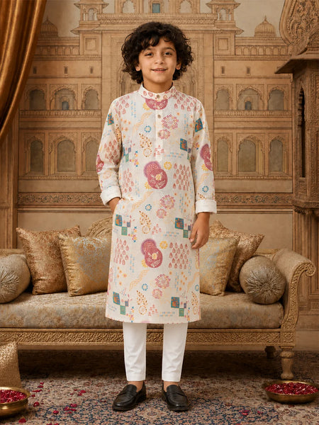 A young boy stands in a decorated traditional room wearing a cream-colored kurta with colorful geometric and floral patterns, paired with white trousers and black loafers.
