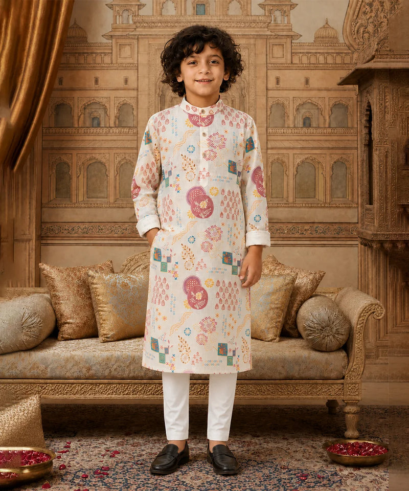 A young boy stands in a decorated traditional room wearing a cream-colored kurta with colorful geometric and floral patterns, paired with white trousers and black loafers.