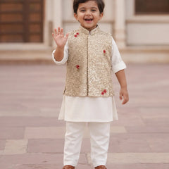 A young boy smiling and waving while wearing a traditional Indian outfit consisting of a white kurta and trousers, a gold sequined vest, and brown sandals, standing outdoors on a stone pavement.