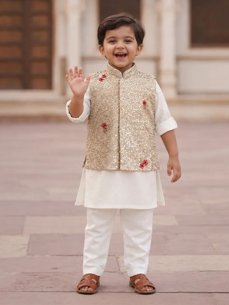A young boy smiling and waving while wearing a traditional Indian outfit consisting of a white kurta and trousers, a gold sequined vest, and brown sandals, standing outdoors on a stone pavement.