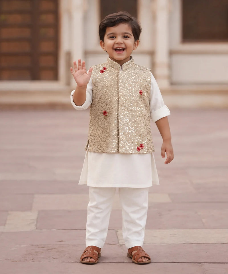 A young boy smiling and waving while wearing a traditional Indian outfit consisting of a white kurta and trousers, a gold sequined vest, and brown sandals, standing outdoors on a stone pavement.